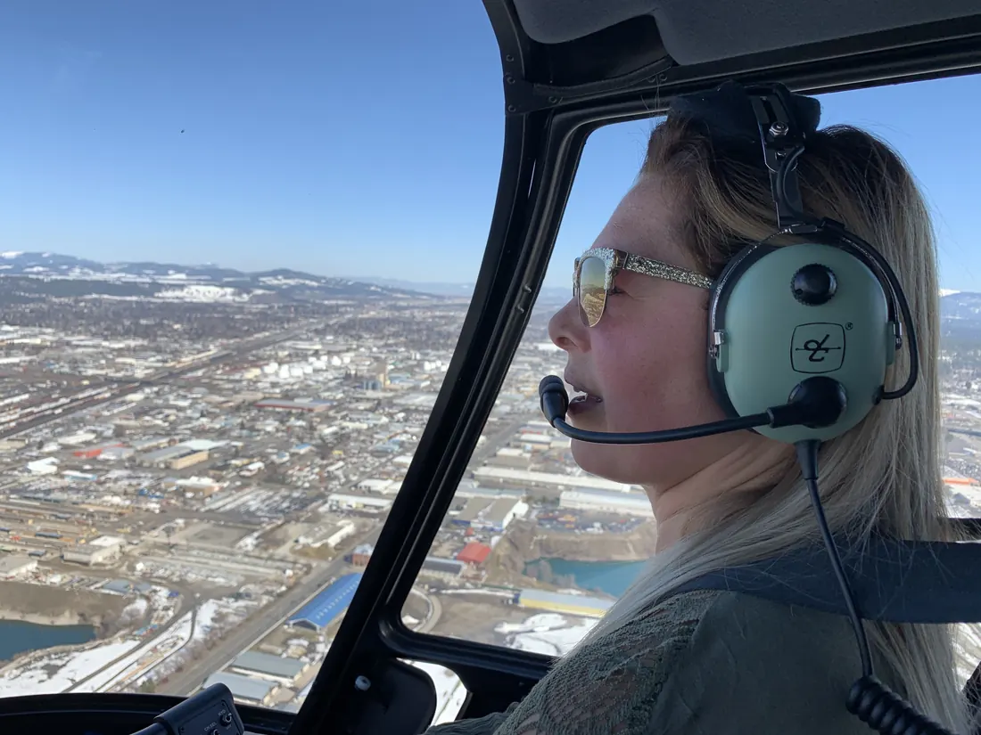 Amy Hartt in a green aviation headset in flight, with a snow-dusted city and hills below.
