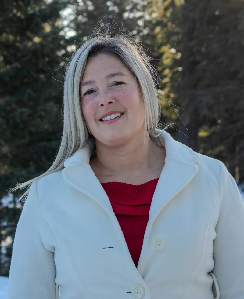 Portrait of Amy Hartt smiling, wearing a cream coat in a snowy pine forest.
